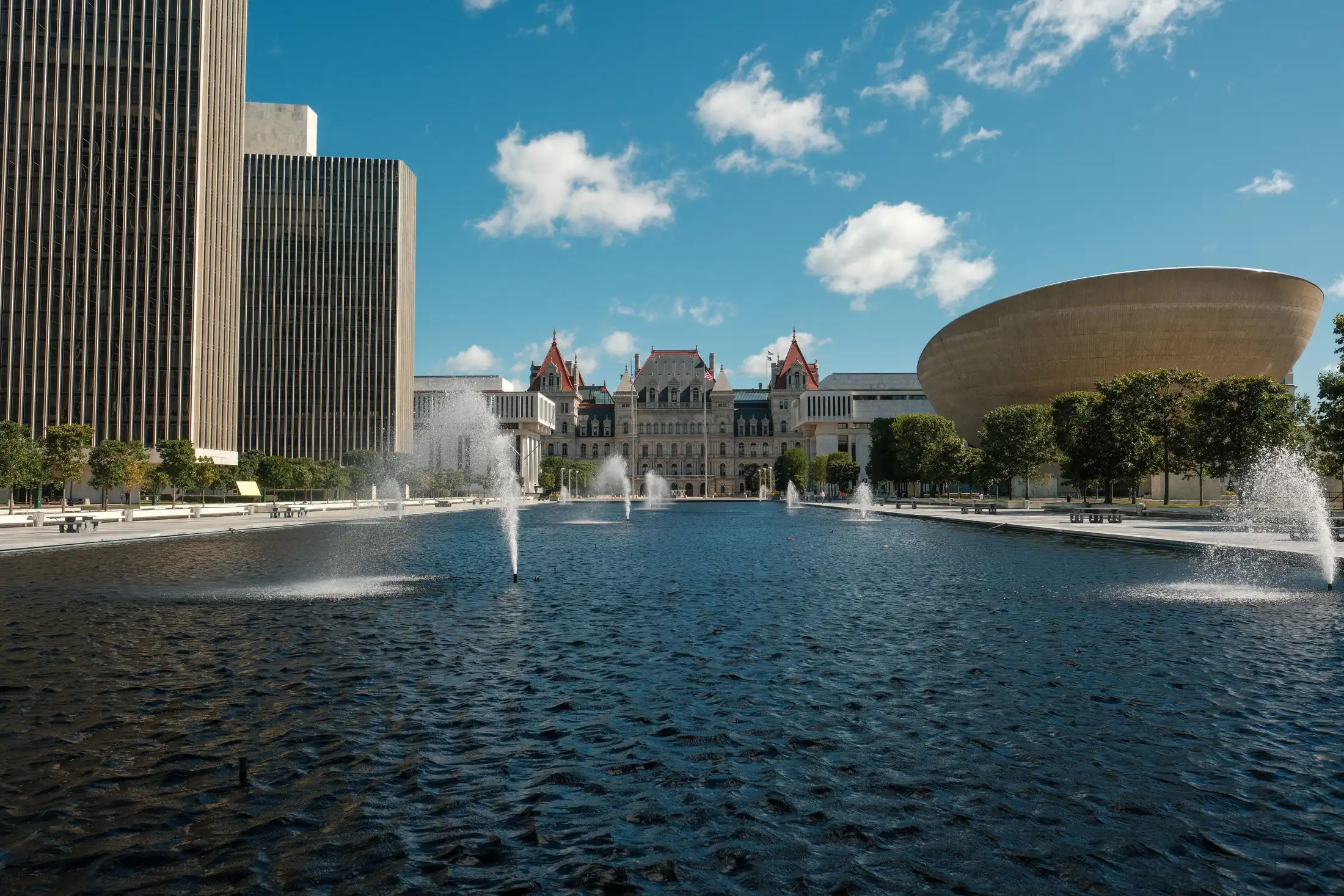 A wide shot of the Empire State Plaza in Albany, New York, on a bright, sunny day. The large reflecting pool with spraying fountains fills the foreground, leading the eye toward the historic New York State Capitol building in the center background. The modern, bowl-shaped building known as The Egg stands on the right, contrasted by tall, vertical skyscraper office buildings on the left under a blue sky with scattered clouds.