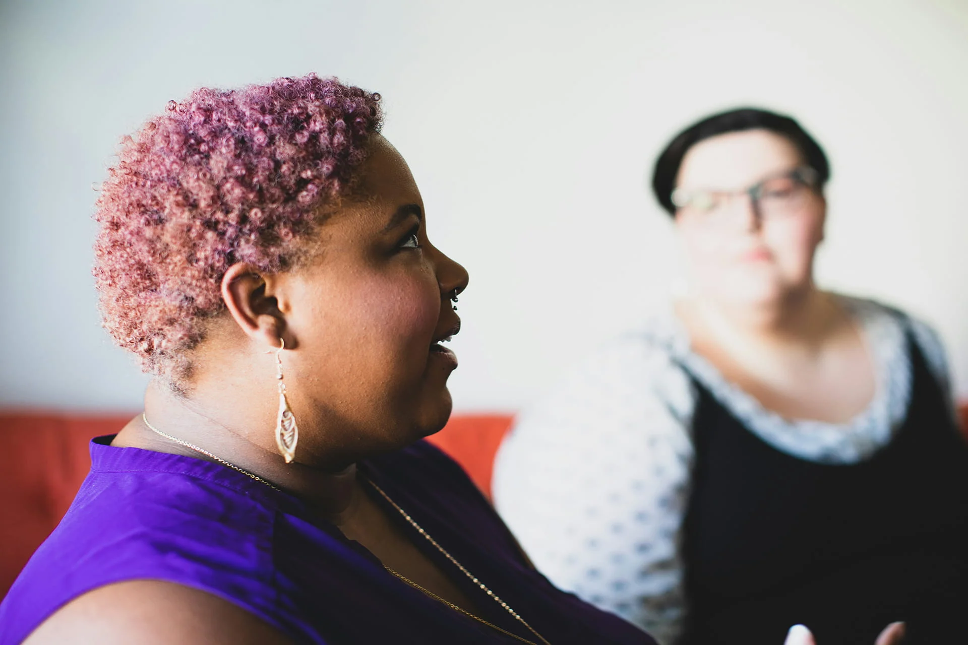 A side profile shot of a person with short, curly pink hair and a purple top, talking while looking upward. Another person wearing glasses sits blurred in the background.