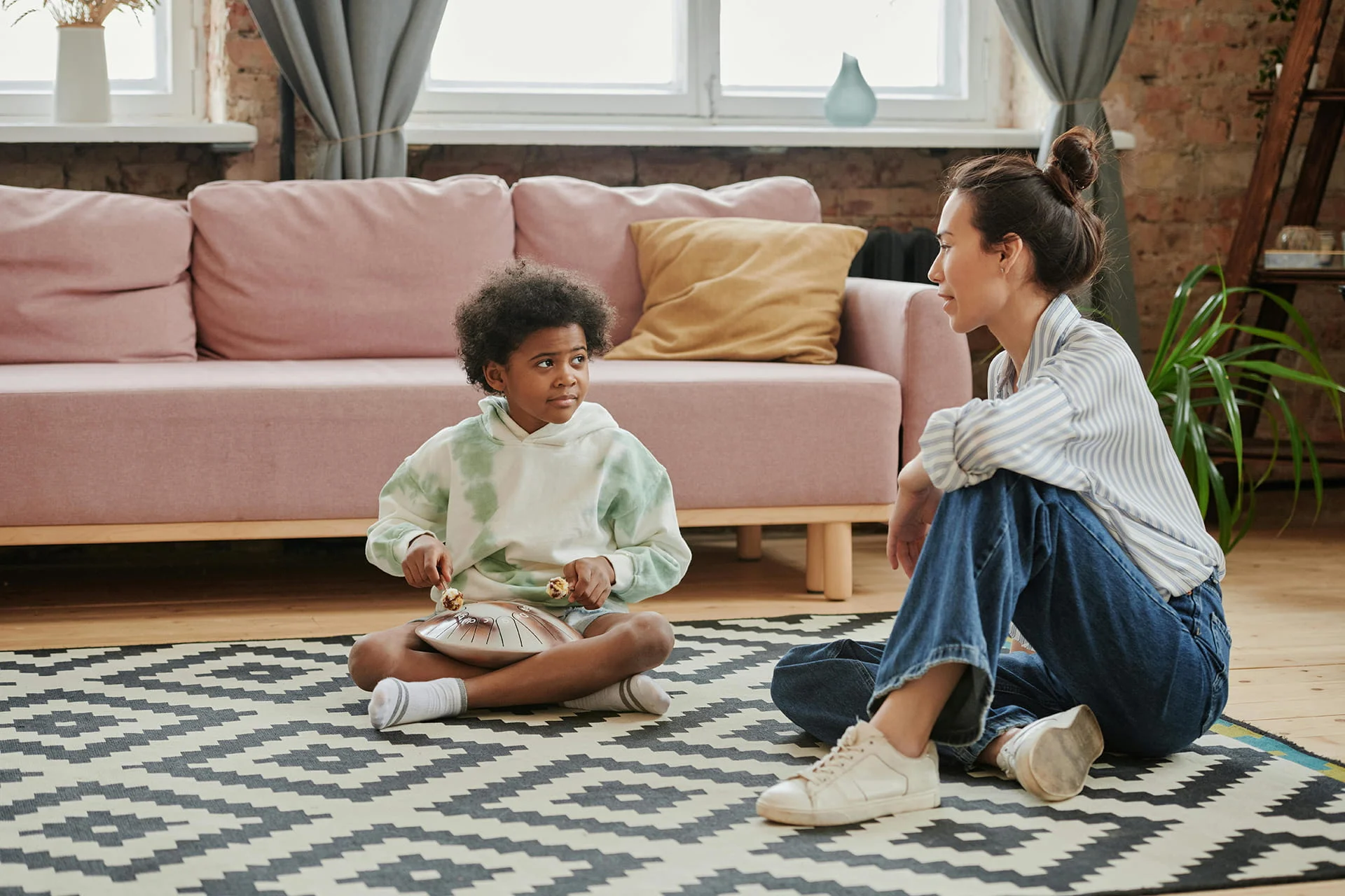 A woman and a young child sit cross-legged on a patterned rug in a living room, engaged in conversation. The child holds mallets over a steel tongue drum in their lap while looking towards the woman, with a pink sofa in the background.
