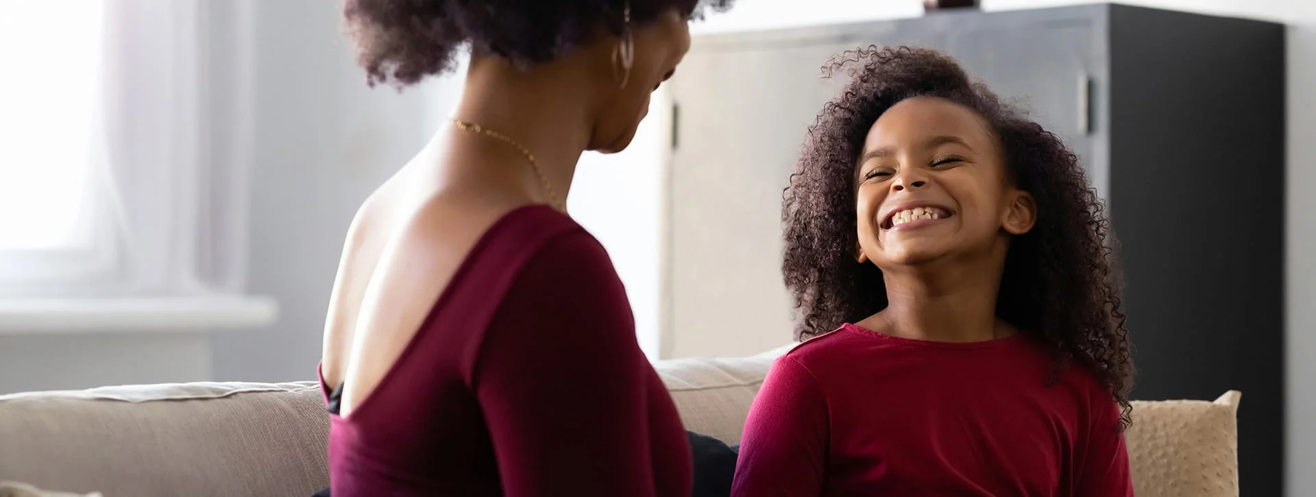 A young girl with curly hair smiles brightly and laughs while sitting on a couch across from a woman, who is seen from a back-profile angle. Both are wearing dark red tops.